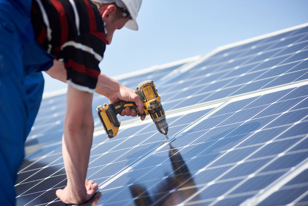 A man with a hard hat on a roof using a screwdriver installing a new solar panel.