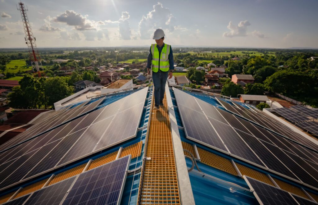 A person in a white hard hat and a yellow safety vest walks on the roof of a building with solar panels.