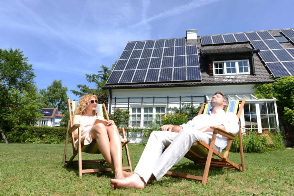 A man and woman relax on chairs on the grass of their backyard with their solar panel roof behind them.